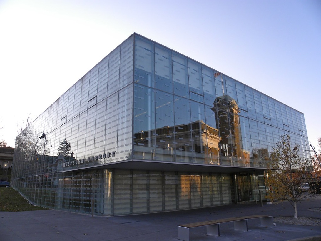 Et moderne, minimalistisk bibliotekbygg med en fasade helt i glass som skaper transparens og slipper inn naturlig lys. Den kubeformede strukturen har et rent, geometrisk design med reflekterende egenskaper som speiler omgivelsene og en historisk bygning i nærheten. Glassfasaden eksemplifiserer moderne arkitektonisk transparens.